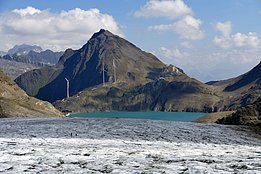 Windräder auf einem Bergkamm hinter einem schmelzenden Gletscher unter blauem Himmel mit Wolken