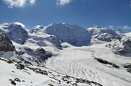 Eine beeindruckende Berglandschaft mit schneebedeckten Gipfeln und Gletschern unter strahlend blauem Himmel. Der Untergrund zeigt Felsen und Schnee, während die massive Bergkette im Hintergrund dominiert.