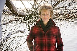 Urte Reckowsky steht am Ufer eines Sees im Winter, trägt eine rote karierte Jacke und schwarze Hose, umgeben von kahlen Bäumen und Schnee.